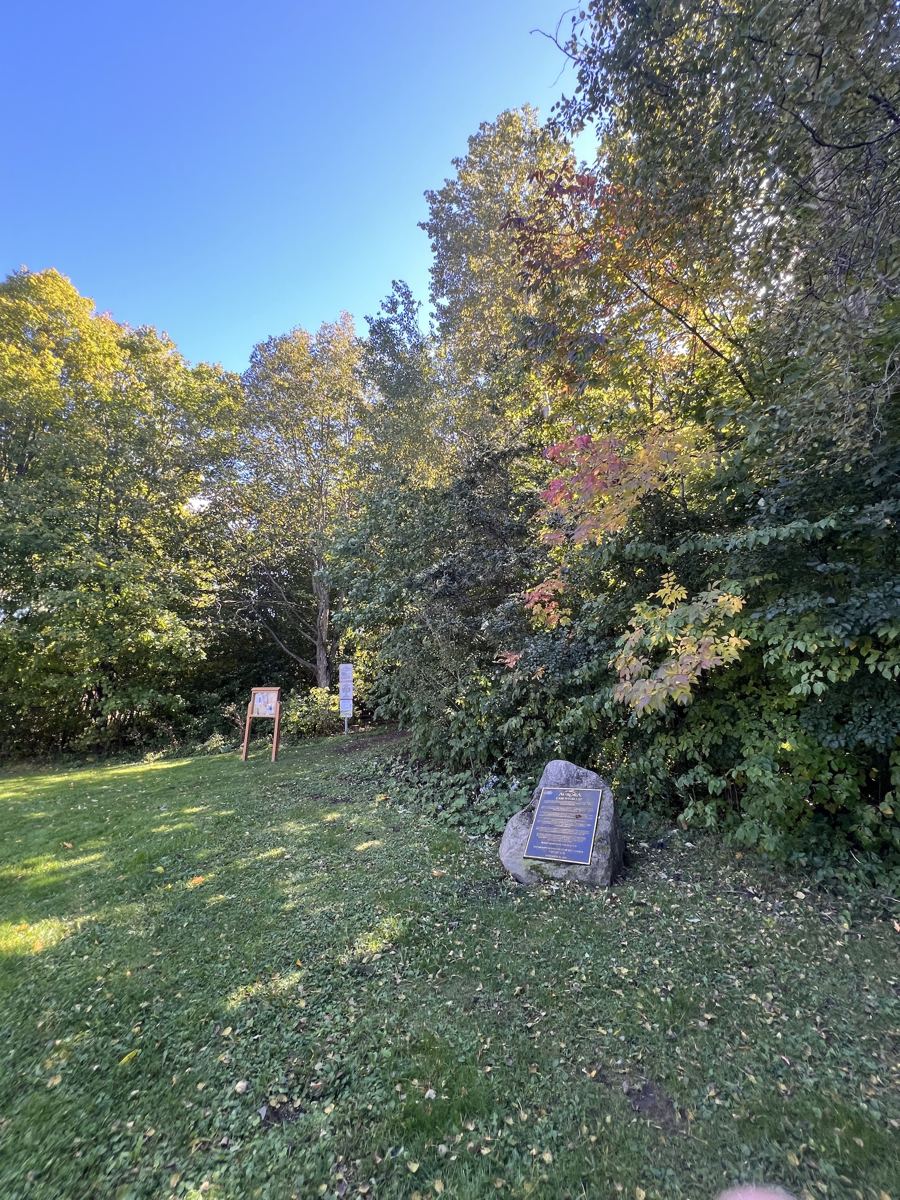 A sunny meadow at the edge of Case Woodlot, with a trail sign, a notice board and a boulder bearing the Case Wood Lot plaque beneath autumn trees