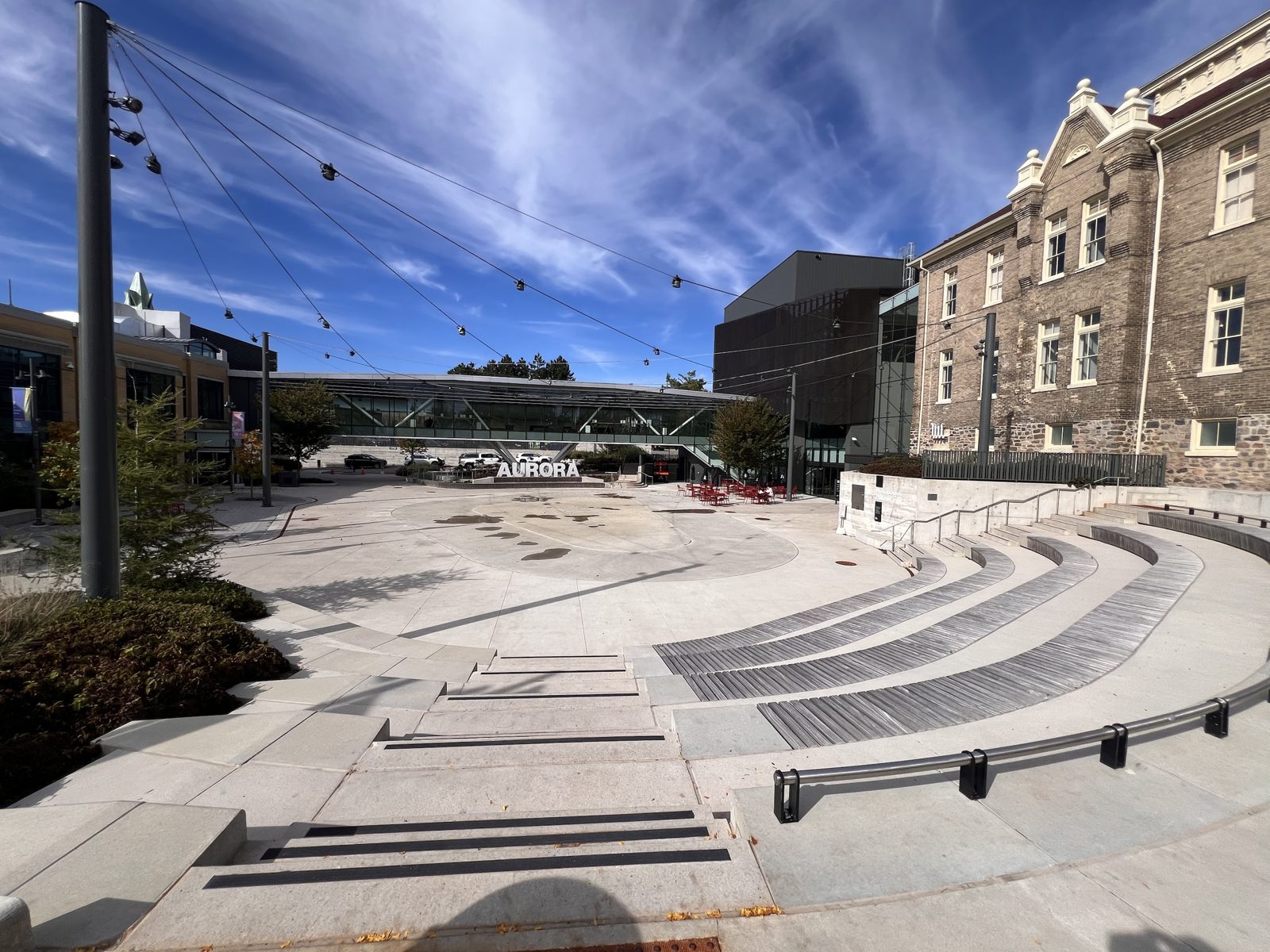 Aurora Town Square: curved amphitheatre steps facing an open plaza, the white AURORA sign, red café chairs and the historic Church Street School
